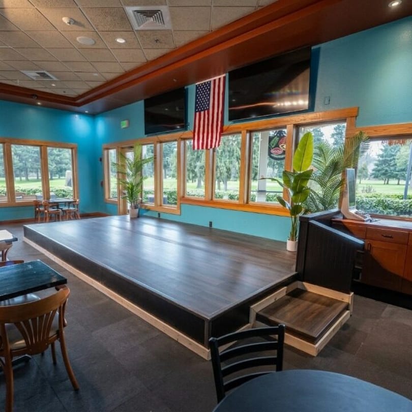 Restaurant interior with a stage, turquoise walls, large windows, and an American flag hanging.
