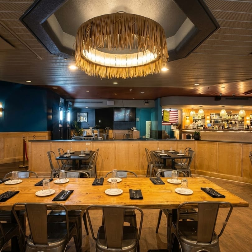 Restaurant interior with a long wooden table in the foreground and a bar in the background.
