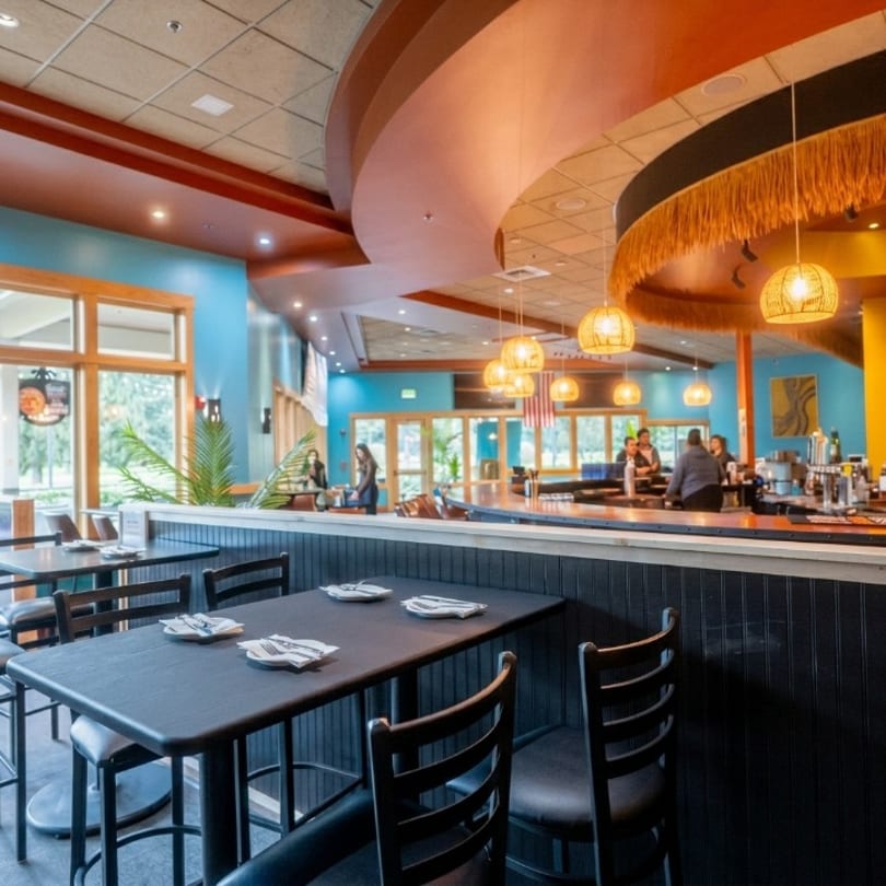 Restaurant interior with blue walls, black tables, and a bar area with people and woven light fixtures.
